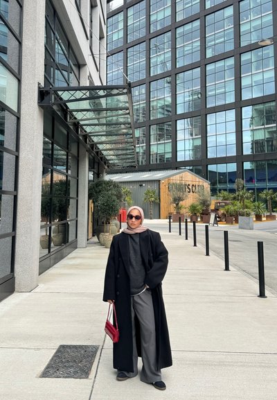Abrigo negro, suéter gris y pantalones anchos, bolso rojo y gafas de sol. De pie en una acera cerca de un edificio con fachada de cristal.