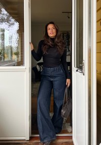 Kobieta o długich włosach, ubrana w czarny golf i szerokie dżinsy, stoi w drzwiach, trzymając brązową torebkę, spoglądając przed siebie.