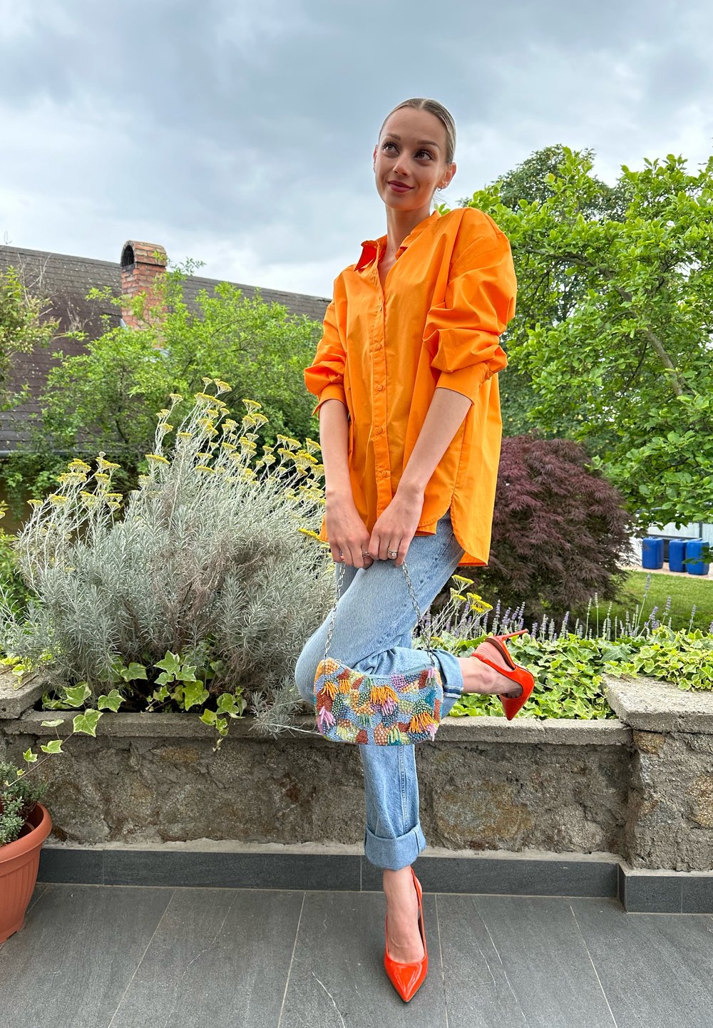 Camisa oversized laranja brilhante, jeans azul claros com uma bolsa colorida de contas e sapatos de salto alto vermelhos. Ambiente externo com vegetação.