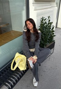 Grauer Strickpullover mit schulterfreiem Design, cremefarbenes Hemd darunter, weite graue Hose, weiße Turnschuhe und eine gelbe Handtasche.