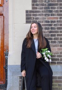 Juodas ilgas paltas ant pilkos palaidinės, juodos kelnės, laiko baltų tulplių ir žalių lapų puokštę, sėdi šalia plytų sienos ir medinių durų.