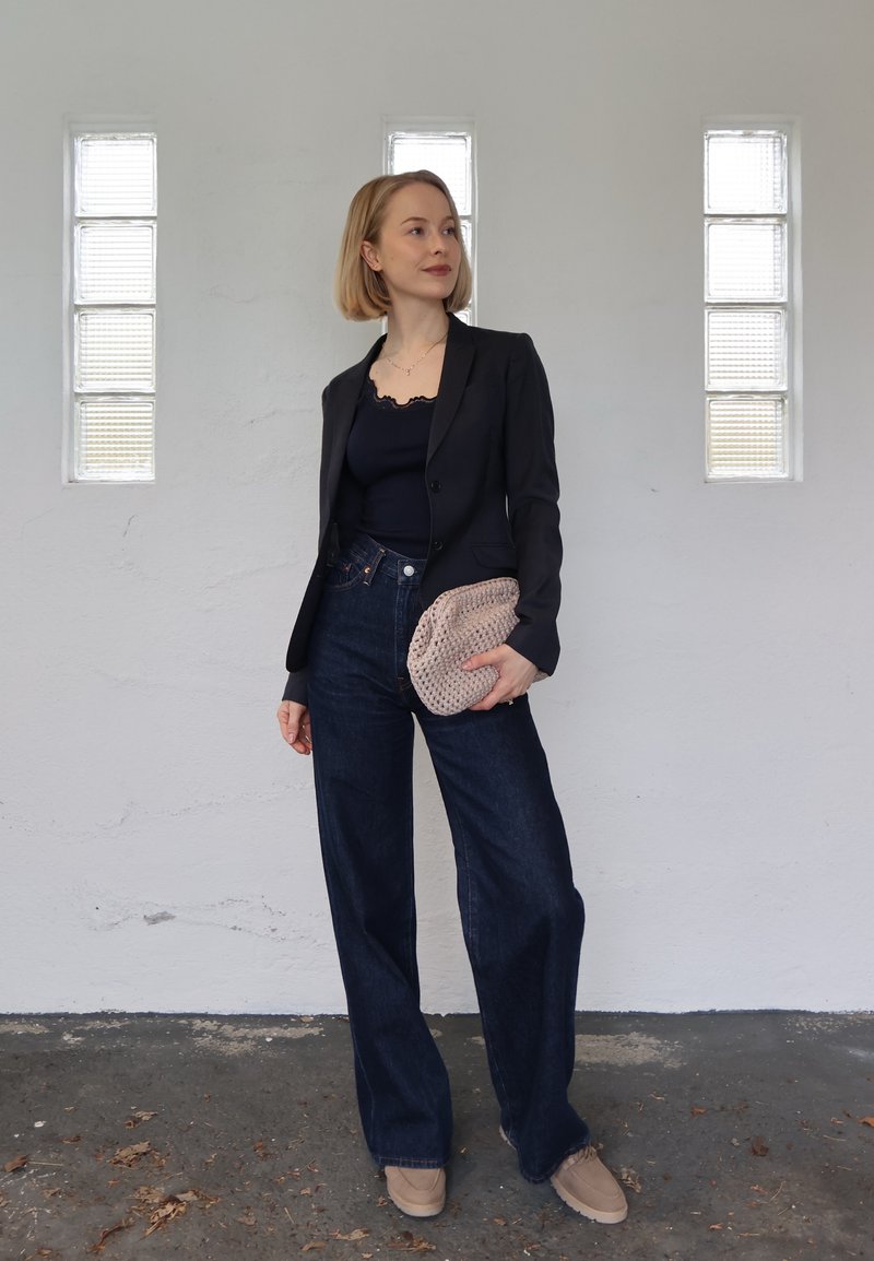 Femme aux cheveux blonds courts, vêtue d'un blazer noir, d'un haut noir, d'un jean large foncé, de chaussures beiges, tenant une pochette en tricot beige.