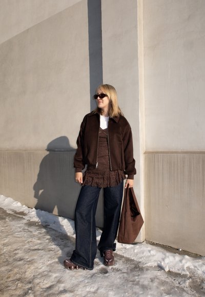 Mujer con chaqueta marrón oscuro, blusa de encaje marrón, vaqueros de pierna ancha y gafas de sol, de pie sobre terreno nevado sosteniendo un bolso marrón a juego.