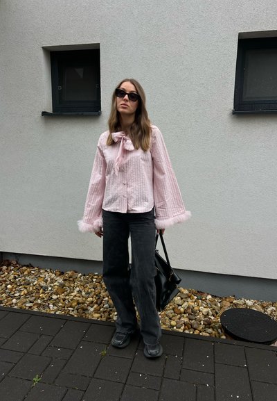 Mujer con blusa rosa texturizada con puños de piel, pantalones negros de pierna ancha, gafas de sol, sosteniendo bolso negro, de pie frente a pared gris con ventanas.