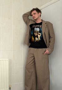 Completo quadrettato marrone con pantaloni larghi e blazer abbinato. Sotto, una t-shirt nera con grafica che raffigura il disegno di una donna.