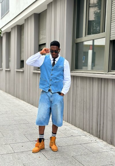 Chaleco de mezclilla en azul claro con botones, camisa blanca, corbata negra, pantalones cortos azul claro y botas marrones. Calcetines a rayas en blanco y negro visibles.