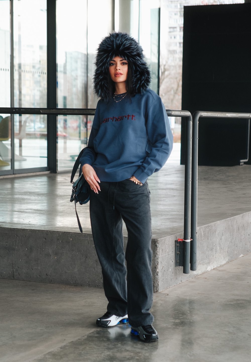 Felpa blu con logo rosso, pantaloni neri e cappuccio foderato in pelliccia nera. Indossa scarpe bianche e blu; tiene una borsa. Sfondo in cemento liscio.