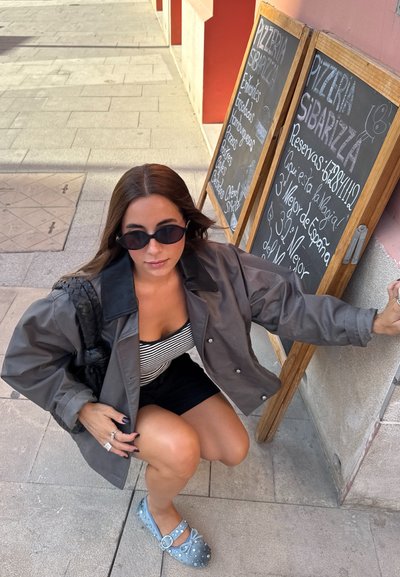 Chaqueta gris de gran tamaño con un cuello negro, camiseta de rayas y pantalones cortos negros. Zapatos azules con adornos. Acera con menús de pizarra.