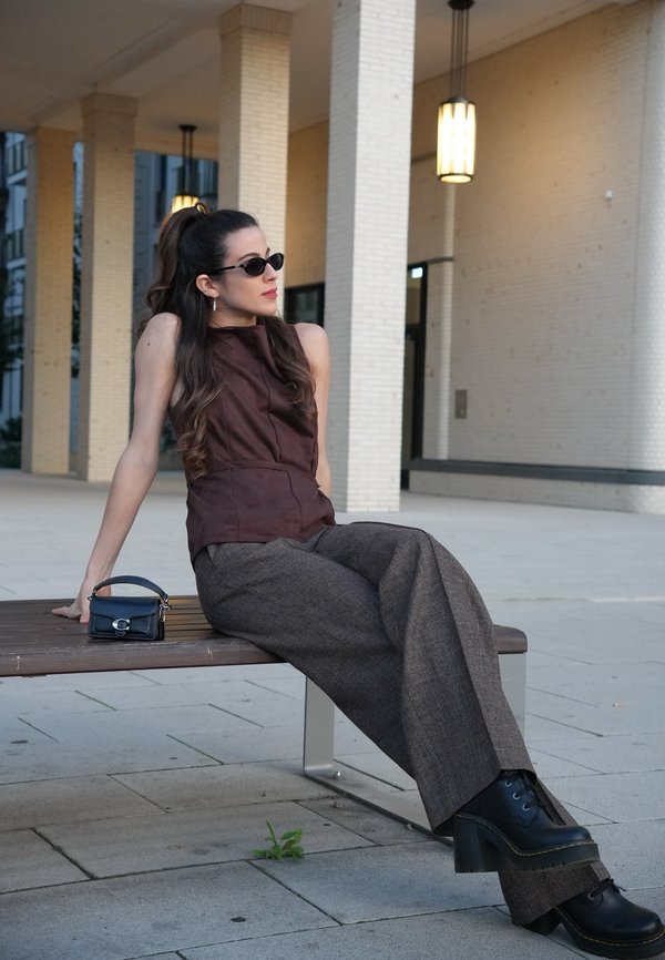 Mujer con gafas de sol, una camiseta sin mangas y pantalones anchos, sentada en un banco al aire libre con un bolso pequeño a su lado.