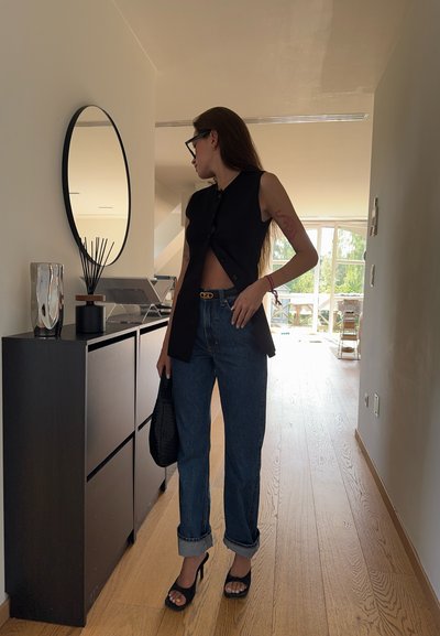 Top negro sin mangas con botones, jeans de mezclilla azul de cintura alta, puños enrollados y sandalias negras de tacón alto, sosteniendo un bolso negro.