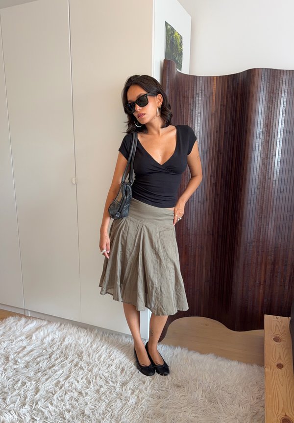Frau trägt schwarzen Top, olivgrünen Rock, schwarze Ballerinas und Sonnenbrille und steht drinnen auf einem weißen Shag-Teppich nahe eines hölzernen Raumteilers.