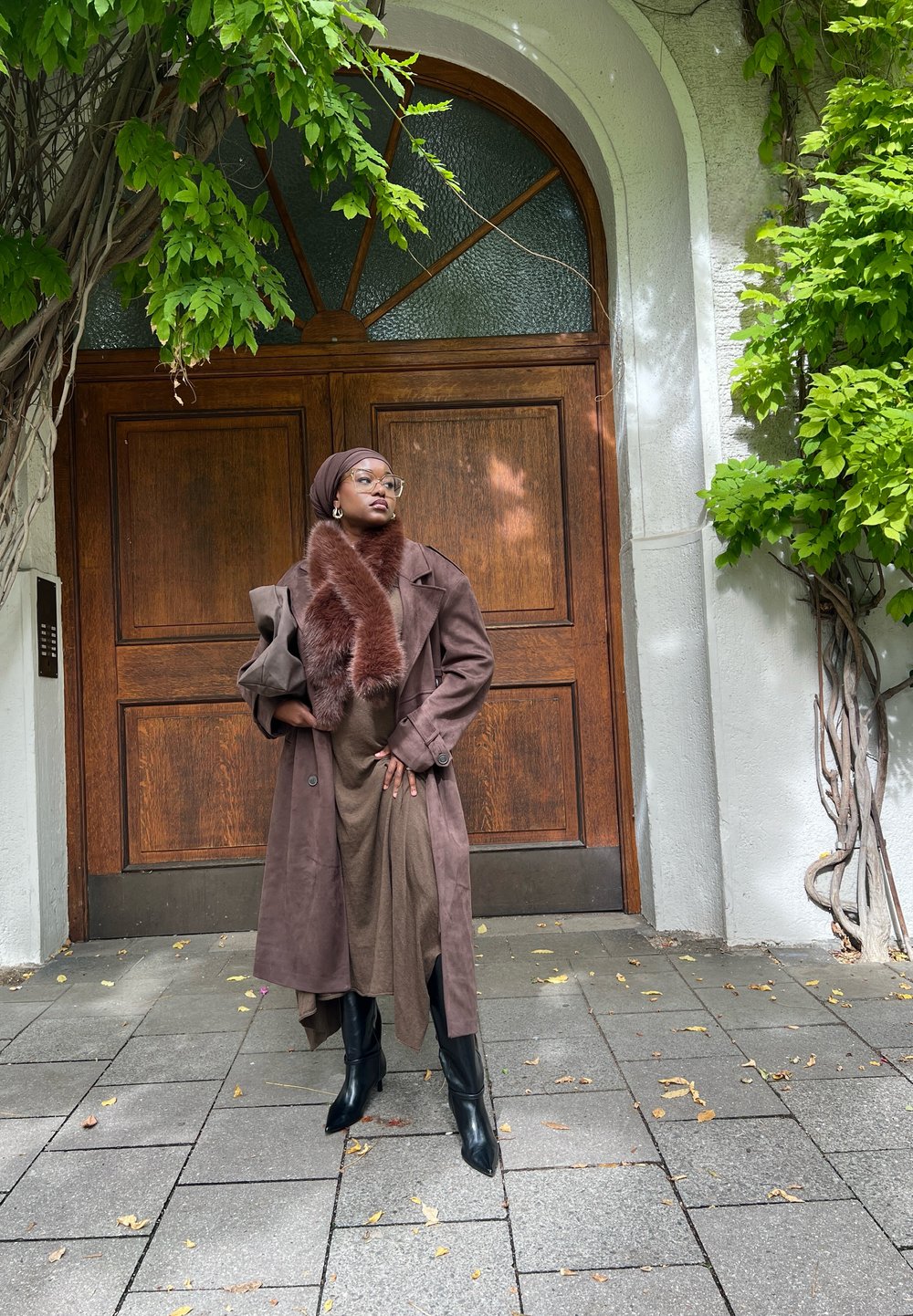 Brun semsket trenchcoat over en brun kjole, komplettert med en faux pels skjerf. Sorte knelengde støvler fullfører antrekket mot en bakgrunn av en tregård.