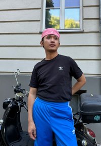 Melns Adidas krekls ar mazu logo, zili treniņbikses ar baltām svītrām un rozā cepure. Stāv blakus melnai skūtera pie sienas.