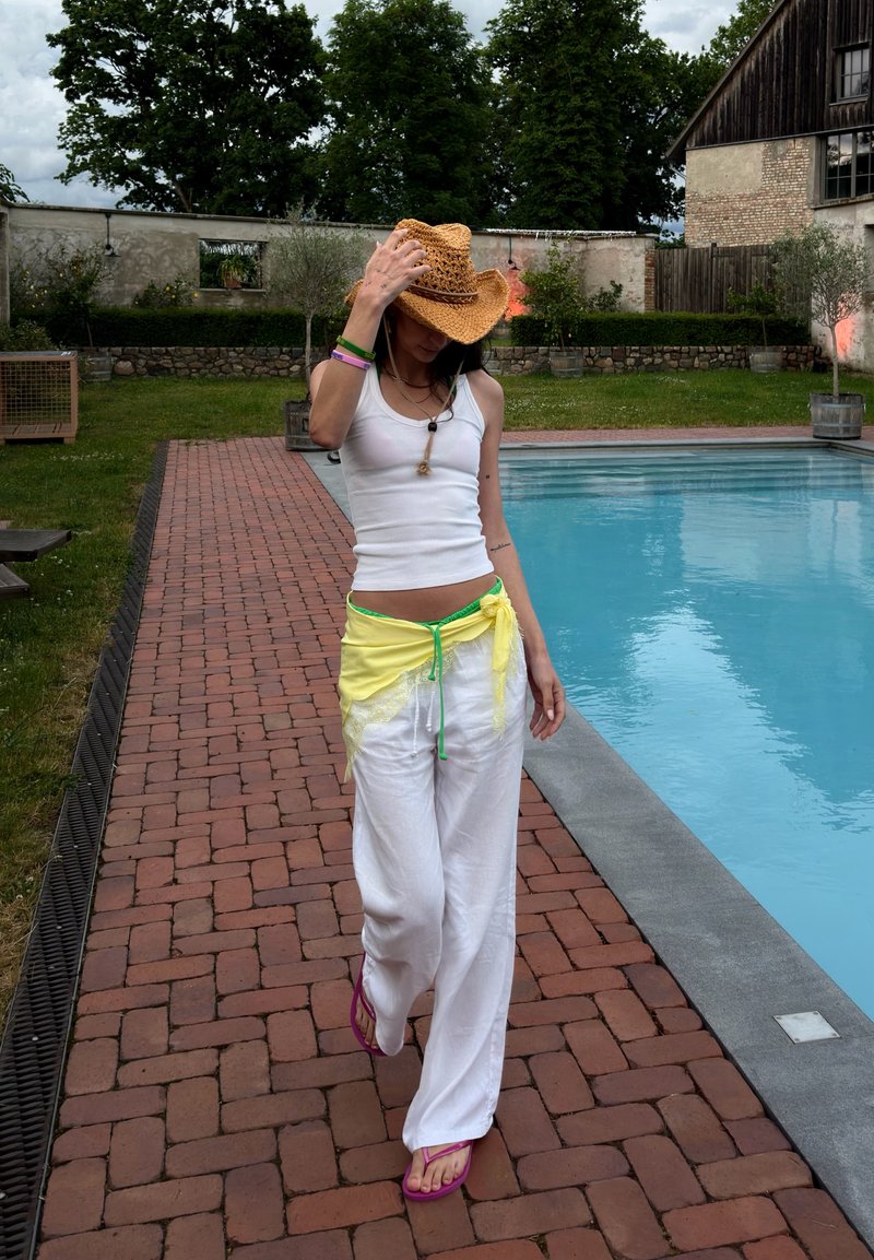 Femme en débardeur blanc et pantalon avec un foulard jaune et un chapeau de paille marchant sur un chemin en briques à côté d'une piscine extérieure.