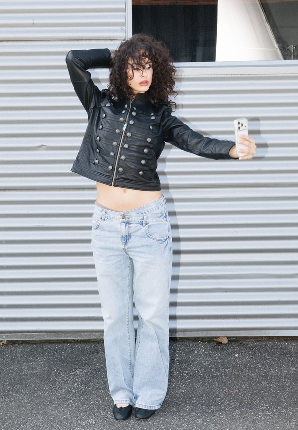 Garbanotais plaukais žmogus juodoje odinėje striukėje ir šviesiuose džinsuose pozuoja, fotografuodamasis prie gofruoto metalo sienos.