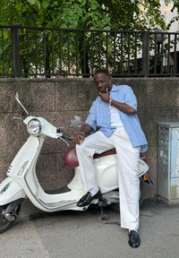 Valkoinen skootteri kromikoreilla, punainen istuin; henkilö, jolla on siniraitainen paita ja valkoiset housut, istuu rennosti. Taustalla vihreitä puita.