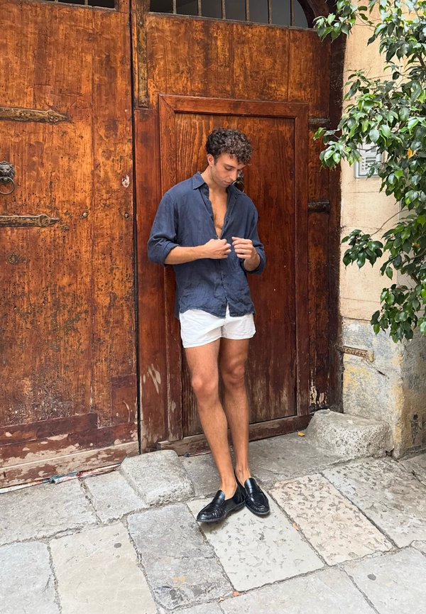 Young man in black loafers, white shorts, and unbuttoned dark blue shirt standing on stone pavement by large wooden door with greenery nearby.