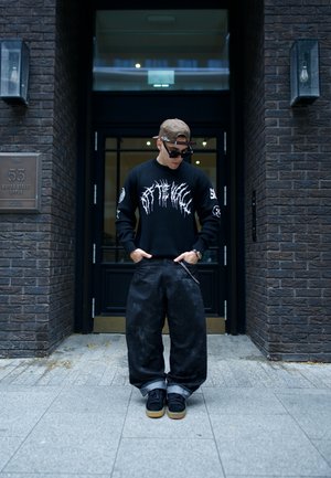 Suéter negro con gráfico blanco, pantalones vaqueros negros holgados y zapatillas negras con suelas de goma. Accesorios incluyen una gorra y gafas de sol.