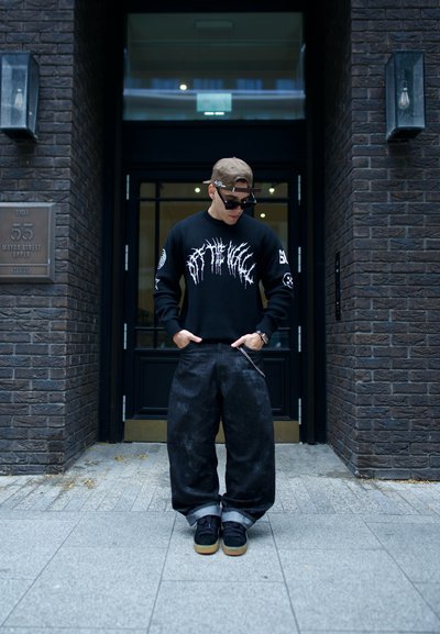 Suéter negro con gráfico blanco, pantalones vaqueros negros holgados y zapatillas negras con suelas de goma. Accesorios incluyen una gorra y gafas de sol.