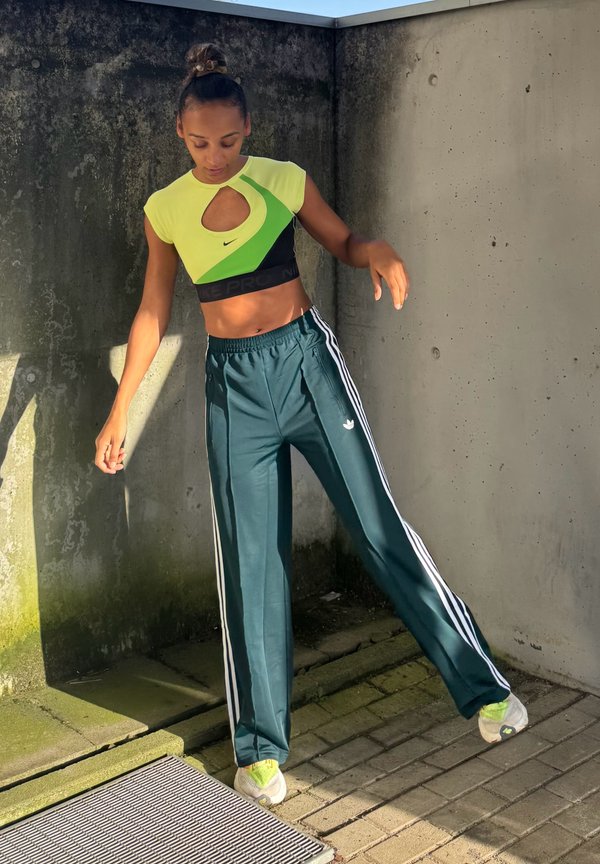 Mujer con top corto verde neón y pantalones deportivos Adidas azul verdoso equilibrándose sobre una pierna al aire libre contra paredes de concreto.