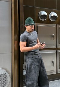 Sivá priliehavá tričko, široké sivé denimové nohavice, zelená čiapka s logom "PORSCHE", opretý o kovovú stenu, sústredený na smartfón.
