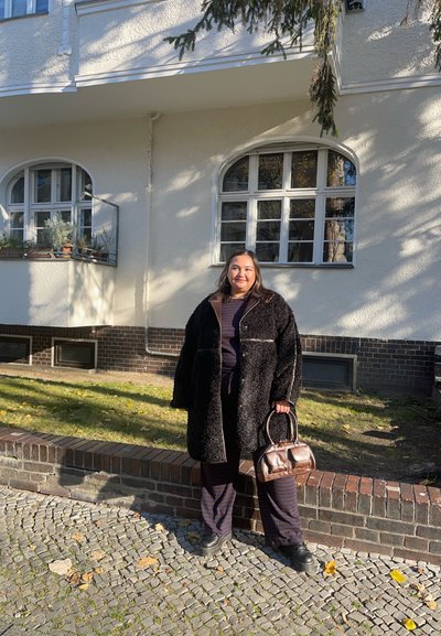Abrigo negro esponjoso con acentos marrones, blusa de rayas azul marino, pantalones a juego y un bolso marrón. Camino de piedra en un entorno al aire libre.