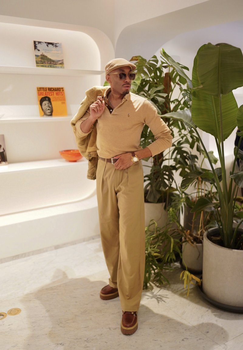 Homme en tenue beige et chapeau pose à l'intérieur près de grandes plantes vertes et d'étagères blanches avec des livres et des objets décoratifs.