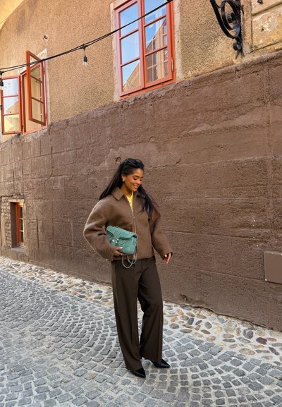 Mujer con cabello largo y oscuro, vestida con una chaqueta y pantalones marrones, sosteniendo un bolso color teal, de pie en una calle empedrada junto a una pared beige.