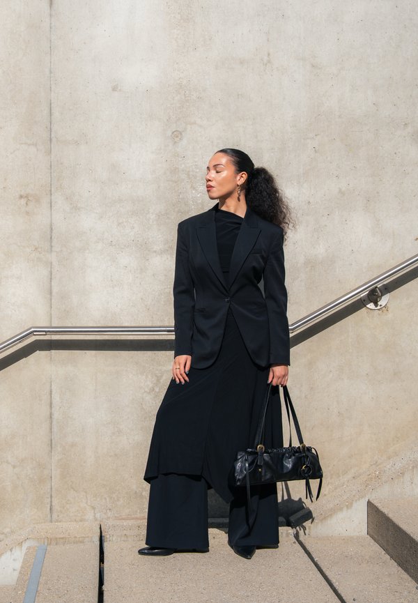 Frau in schwarzem Blazer und weitem Hosenanzug, die eine schwarze Handtasche hält und auf einer Treppe vor einer Betonwand mit Metallgeländer steht.
