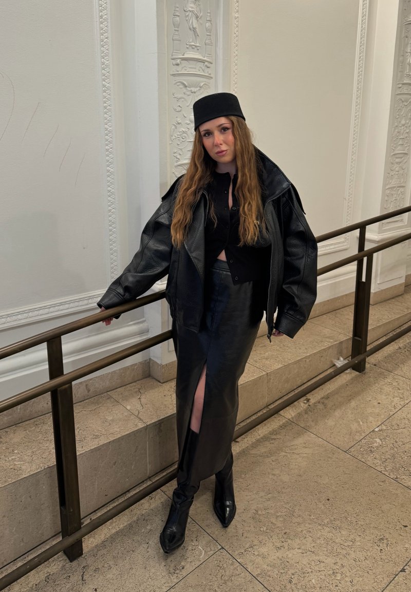 Femme aux longs cheveux portant une veste en cuir noire, une jupe fendue à la cuisse, des bottes et un chapeau, appuyée contre une rambarde métallique dans un espace intérieur orné.