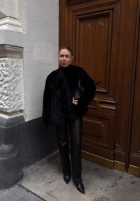 Kvinde iført en sort pelsfrakke, sorte læderbukser og spidse sorte støvler, holdende en sort håndtaske, stående ved en trædør.