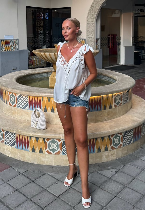 Une femme vêtue d’un chemisier blanc, d’un short en jean et de talons se tient près d’une fontaine carrelée dans une cour couverte.