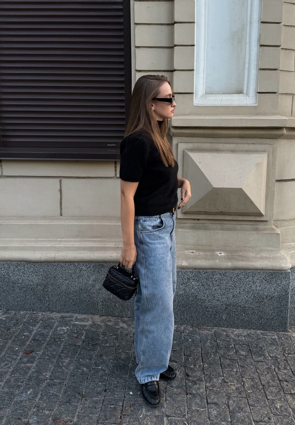 Mujer con top negro y gafas de sol, sosteniendo un pequeño bolso negro, de pie en una calle empedrada frente a una pared de piedra beige de un edificio.