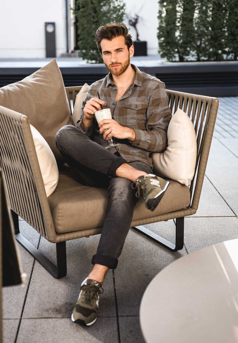 Gevoerd stoeltje met een beige stof, een jonge man in een grijs geruit overhemd houdt een milkshake vast. Hij draagt donkere spijkerbroek en olijfgroene sneakers.