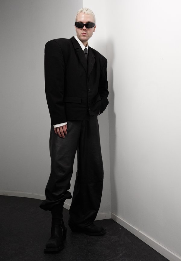 Person med kort blondt hår og svarte solbriller iført oversized svart blazer, løse svarte bukser og svarte støvler, stående mot en hvit vegg.