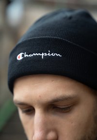 Gorro de punto negro con el logotipo "Champion" en cursiva blanca y un pequeño detalle rojo. Vista en primer plano que resalta la textura y los detalles de la costura.