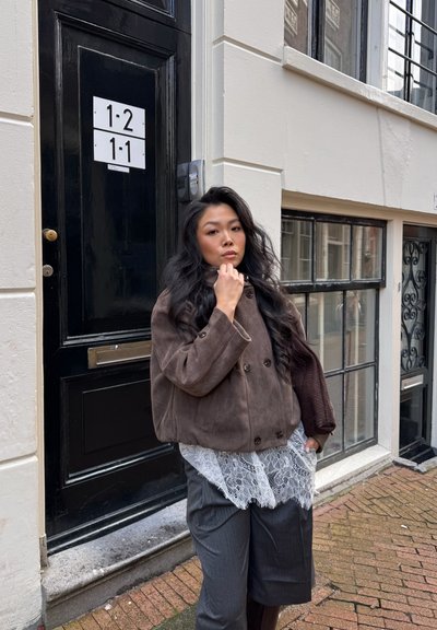 Joven mujer con cabello largo y oscuro que lleva una chaqueta marrón, top de encaje y pantalones grises, está de pie en una calle de ladrillos cerca de una puerta y ventanas negras.