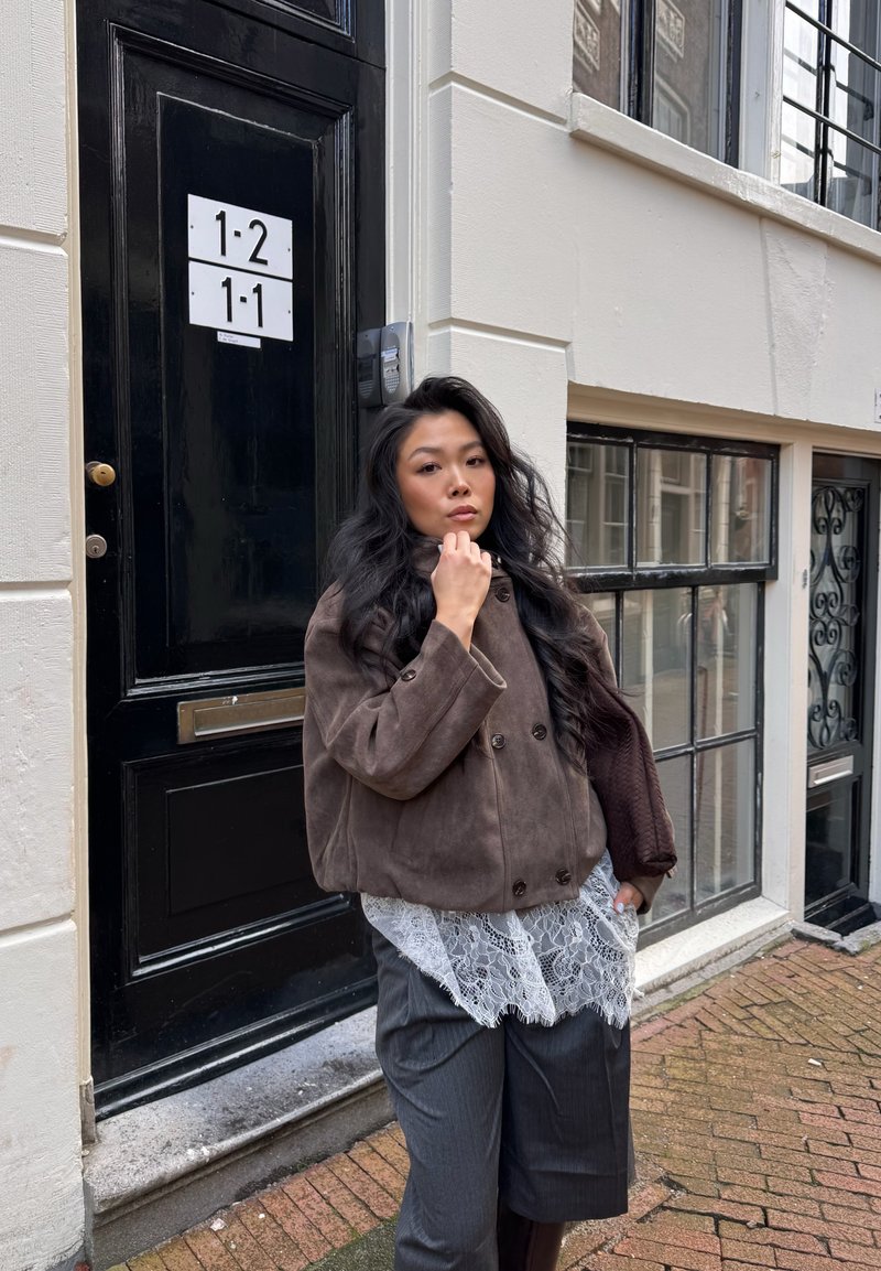 Jeune femme aux longs cheveux foncés portant une veste marron, un haut en dentelle et un pantalon gris, debout dans une rue pavée près d'une porte et de fenêtres noires.