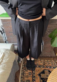 Pantalon large noir à jambe droite, taille haute, tissu lisse et plis marqués ; associé à des baskets noires et des chaussettes blanches.