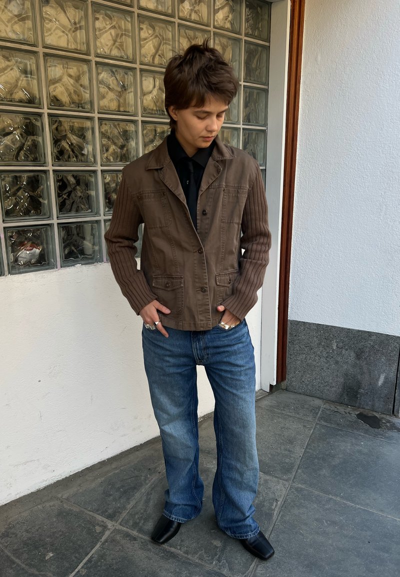 Personne regardant vers le bas portant une veste marron avec des manches tricotées, un jean bleu, des chaussures noires et des bagues argentées, debout sur un sol carrelé à côté d'un mur en blocs de verre.