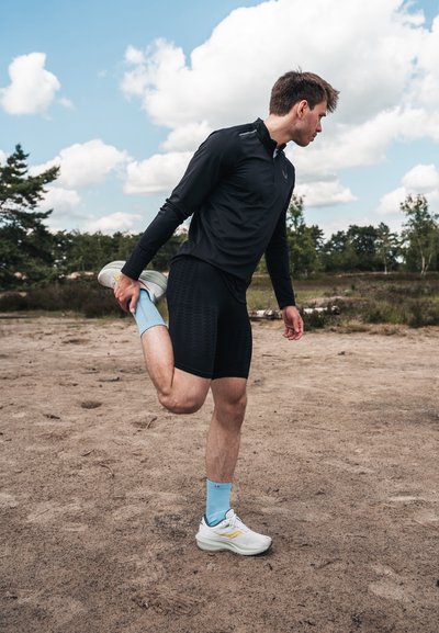 Top negro de manga larga, pantalones cortos negros, calcetines azules y zapatillas deportivas blancas. La figura estira una pierna sobre un terreno arenoso frente a un cielo nublado.