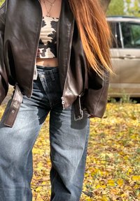 Persona con capelli lunghi rossi indossa un top a strisce bianche e nere, una giacca di pelle marrone e jeans blu larghi, in piedi su foglie autunnali accanto a un veicolo.