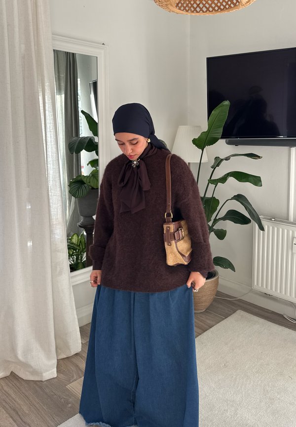 Nő fekete fejkendőben, barna pulóverben és hosszú kék szoknyában, bent áll egy tükör, növény és televízió közelében.