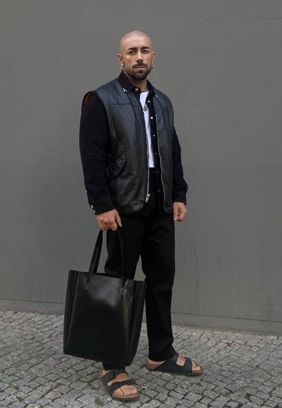 Chaleco de cuero negro sobre una camisa de manga larga negra, pantalones negros y sandalias. Sostiene un gran bolso tote de cuero negro. Fondo liso.