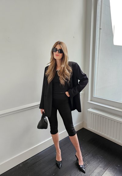 Blazer negro oversize sobre una camiseta negra, combinado con leggings cortos negros y tacones puntiagudos negros. Los accesorios incluyen un bolso de mano negro trenzado.