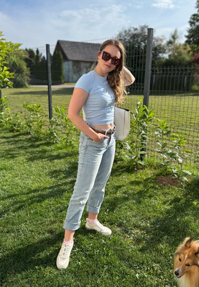 Camiseta azul claro recortada de mangas cortas, jeans de lavado claro con puños, zapatillas blancas y gafas de sol de gran tamaño; fondo de hierba verde.