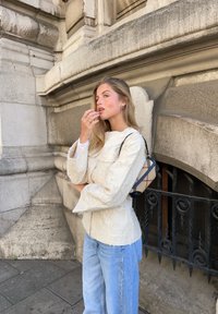 Casaco creme texturizado com fecho éclair e dois bolsos no peito, combinado com jeans de perna larga azul clara. Ela leva uma pequena bolsa de ombro com padrão.
