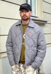Casaco cinza acolchoado com gola e fecho éclair, usado sobre uma camisa verde-oliva. Apresenta um logótipo vermelho e calças estampadas.
