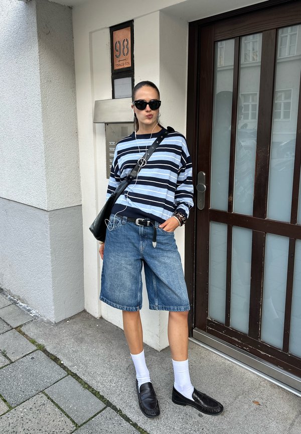 Persona con gafas de sol, camiseta de manga larga a rayas, pantalones cortos vaqueros, calcetines blancos y zapatos negros, de pie junto a la entrada del edificio con puerta y placa de dirección.