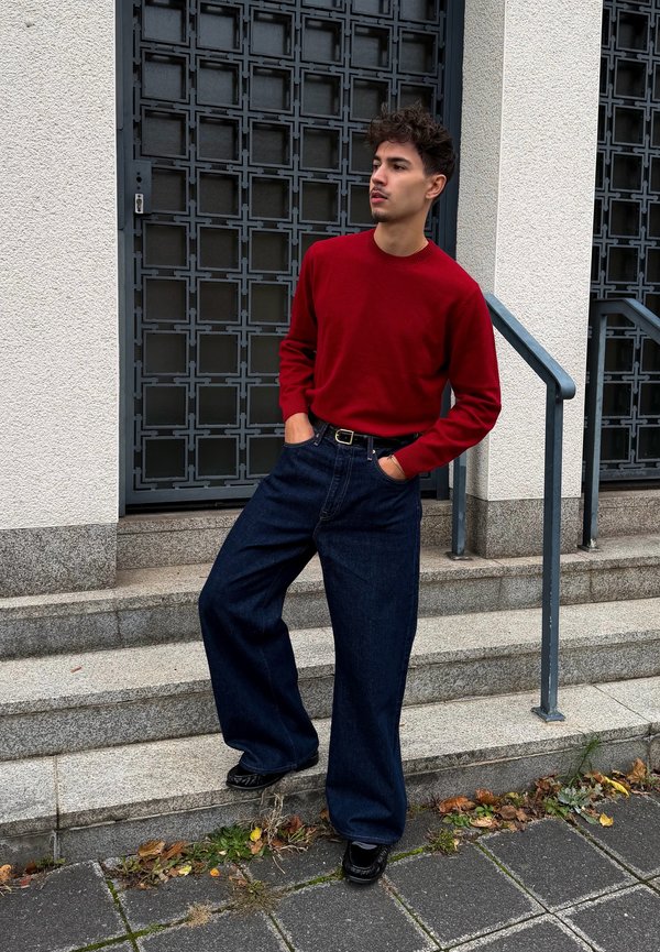 Un jeune homme portant un pull rouge et un jean à jambes larges s’appuie contre une rampe dans un escalier en pierre, à l’extérieur d’un bâtiment.